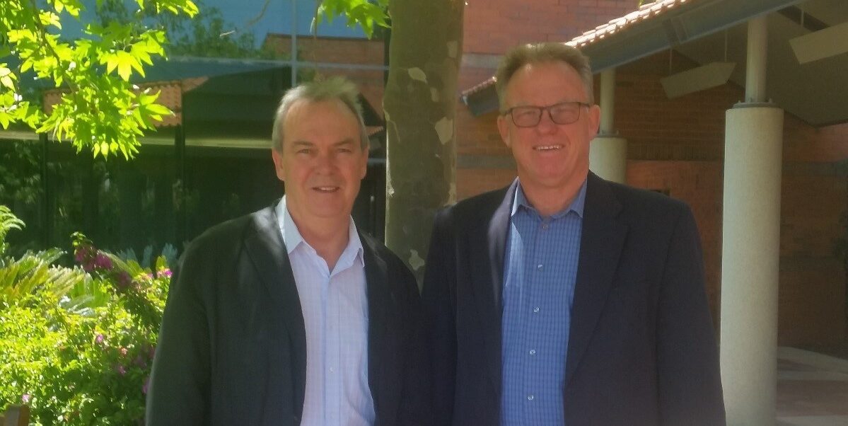 Professor John Phillimore and Dr Martin Whitely are both wearing blue button up shirts and dark jackets. They are standing on Curtin Perth campus in front of a tree, smiling at the camera.