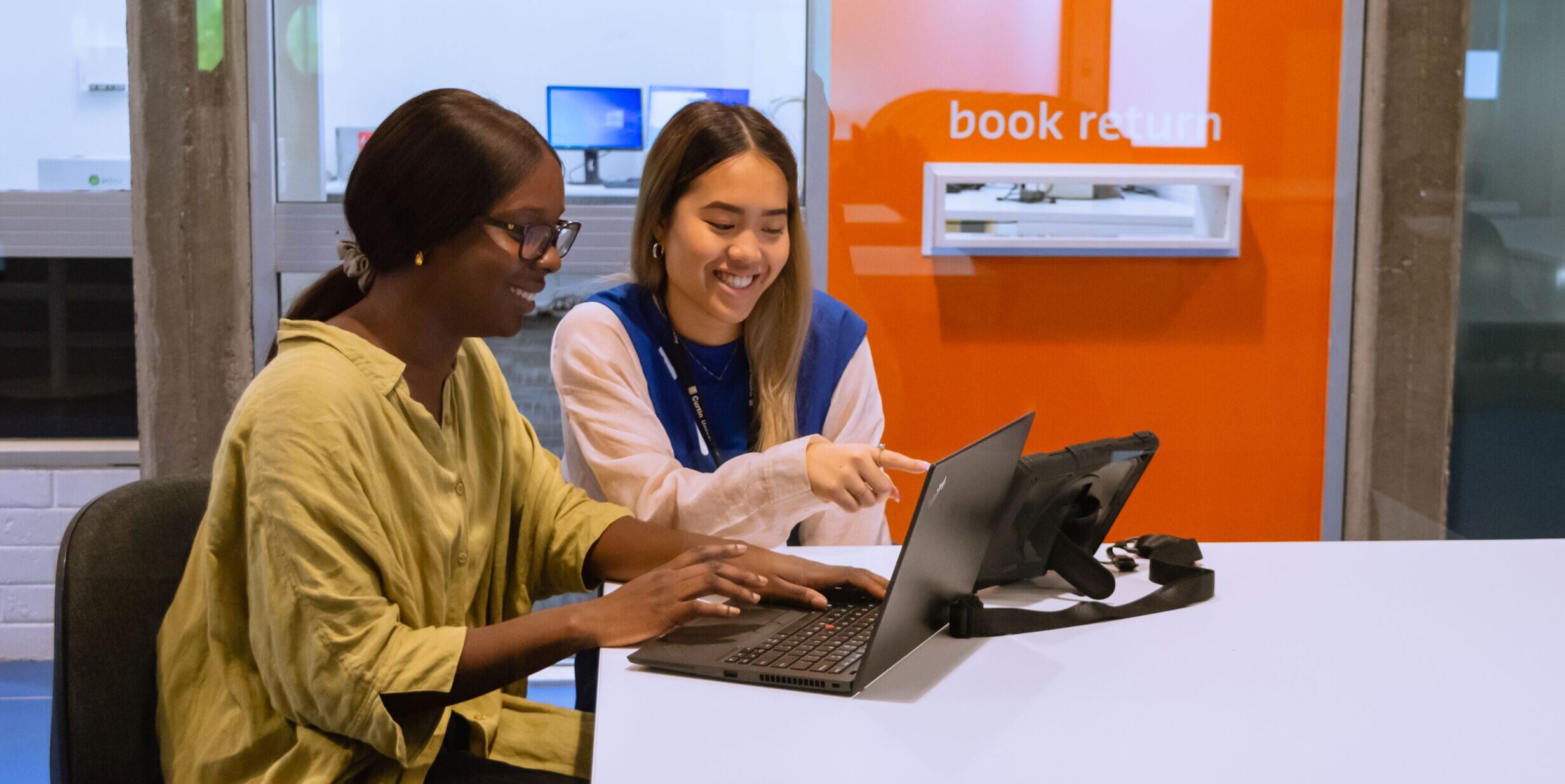 A library staff member helps a student sitting at a desk with their laptop