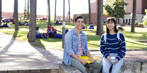 Two students sit outside smiling at the camera, with pinetrees and students sitting on the grass behind them