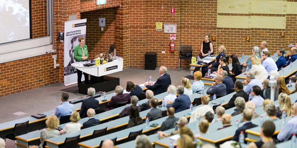 Rows of people sit and listen to University librarian Catherine Clark who is speaking at the lecture podium. She wears a green jacket and glasses.