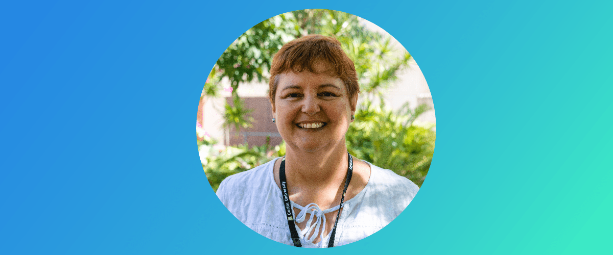 Jill wears a white blouse and Curtin lanyard. She has orange hair is standing outside smiling at the camera in front of trees and dappled light.