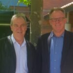 Professor John Phillimore and Dr Martin Whitely are both wearing blue button up shirts and dark jackets. They are standing on Curtin Perth campus in front of a tree, smiling at the camera.