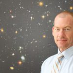 Steven Tingay wears a white shirt and striped tie. He is in front of a poster of the universe.