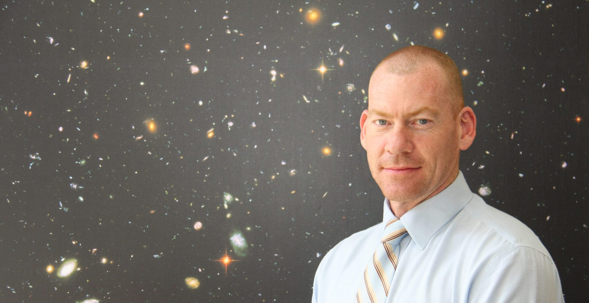 Steven Tingay wears a white shirt and striped tie. He is in front of a poster of the universe.