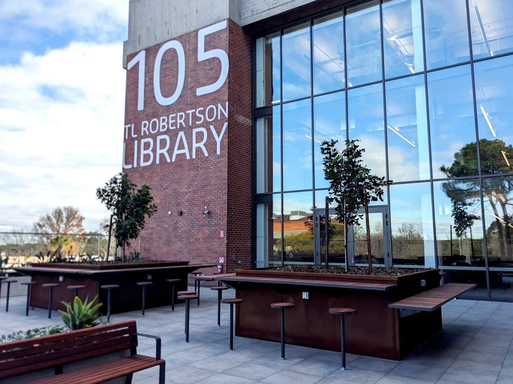 Level 4 outdoor terrace. Wooden benches, some trees and the TL Robertson Library sign is visible on the side of the building.